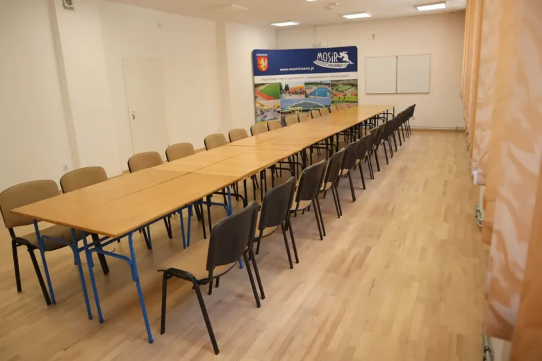 Sala z długim stołem i wieloma krzesłami, w tle logo mosir